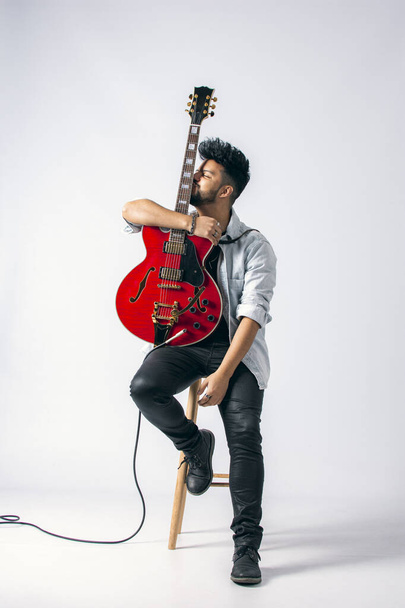 Studio shot of a brown-skinned Latino man with dark hair and beard sitting on a wooden stool wearing a jeans jacket and holding a red electric guitar on a white background. - 写真・画像