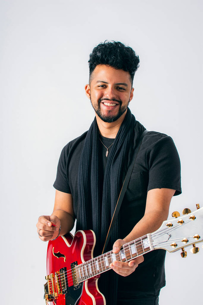 Studioaufnahme eines braunhäutigen Latinos mit dunklen Haaren und Bart in schwarzer Kleidung, der lachend eine rote E-Gitarre auf weißem Hintergrund spielt. - Foto, Bild