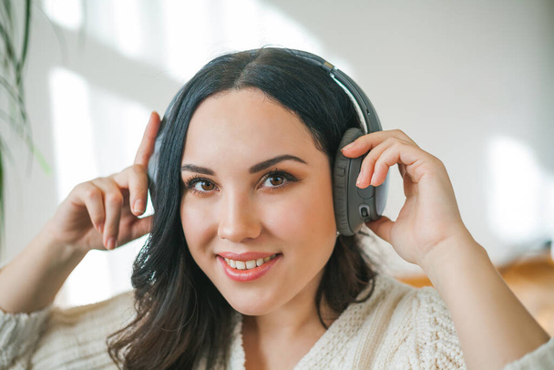 Young beautiful dark-haired curly woman in a knitted cardigan listens to music on headphones while sitting in an armchair in a cozy home. Rest. - Photo, Image