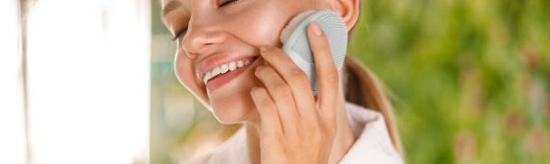 Retrato recortado de una mujer joven que usa albornoz sonriendo mientras usa un cepillo facial de silicona para limpiar la piel - Foto, Imagen