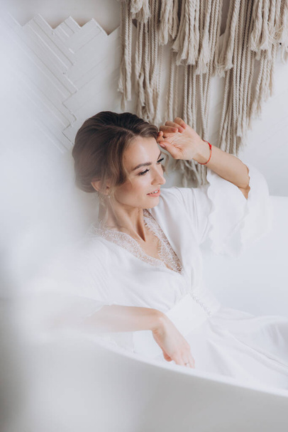 Morning of the bride in a rustic style, in a white bathrobe and a white bouquet on the bed and in the bathroom by the window in a photo studio, in a hotel - 写真・画像