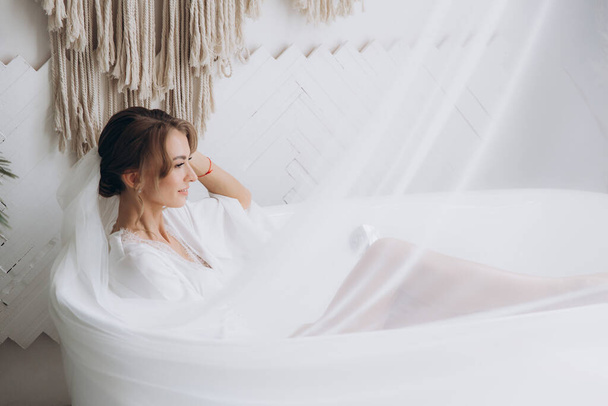 Manhã da noiva em estilo rústico, em roupão branco e um buquê branco na cama e no banheiro pela janela em um estúdio de fotos, em um hotel - Foto, Imagem