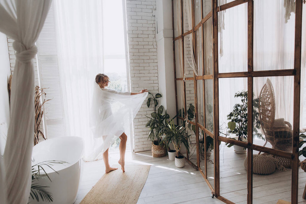 Morsiamen aamu maalaistyyliin, valkoisessa kylpytakissa ja valkoisessa kukkakimpussa sängyllä ja kylpyhuoneessa ikkunassa valokuvaamossa, hotellissa - Valokuva, kuva