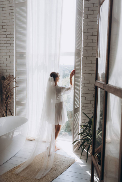 Morning of the bride in a rustic style, in a white bathrobe and a white bouquet on the bed and in the bathroom by the window in a photo studio, in a hotel - Foto, Imagem