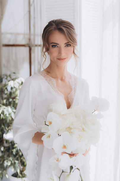 Morning of the bride in a rustic style, in a white bathrobe and a white bouquet on the bed and in the bathroom by the window in a photo studio, in a hotel - Fotó, kép