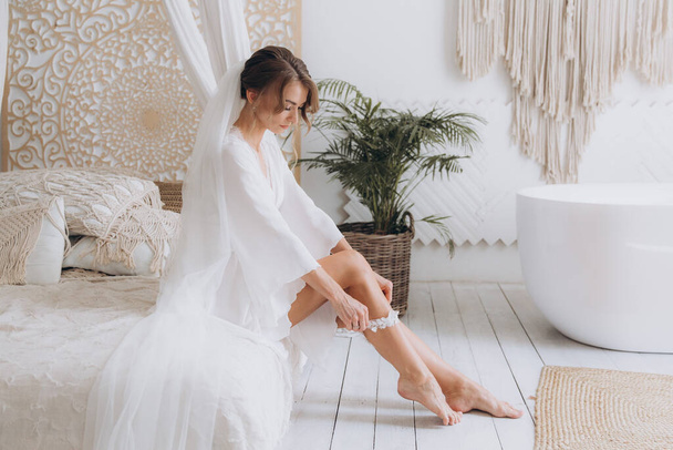 Mañana de la novia en un estilo rústico, en un albornoz blanco y un ramo blanco en la cama y en el baño por la ventana en un estudio de fotografía, en un hotel - Foto, imagen