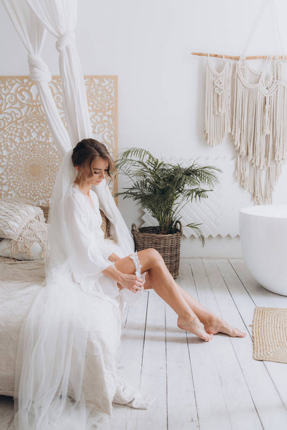 Ochtend van de bruid in een rustieke stijl, in een witte badjas en een wit boeket op het bed en in de badkamer door het raam in een fotostudio, in een hotel - Foto, afbeelding