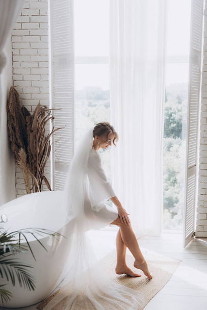 Morning of the bride in a rustic style, in a white bathrobe and a white bouquet on the bed and in the bathroom by the window in a photo studio, in a hotel - Foto, afbeelding