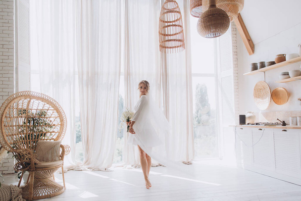 Ochtend van de bruid in een rustieke stijl, in een witte badjas en een wit boeket op het bed en in de badkamer door het raam in een fotostudio, in een hotel - Foto, afbeelding