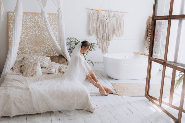A menyasszony reggele rusztikus stílusban, fehér fürdőköpenyben és fehér csokorban az ágyon és a fürdőszobában az ablak mellett egy fotóstúdióban, egy hotelben - Fotó, kép
