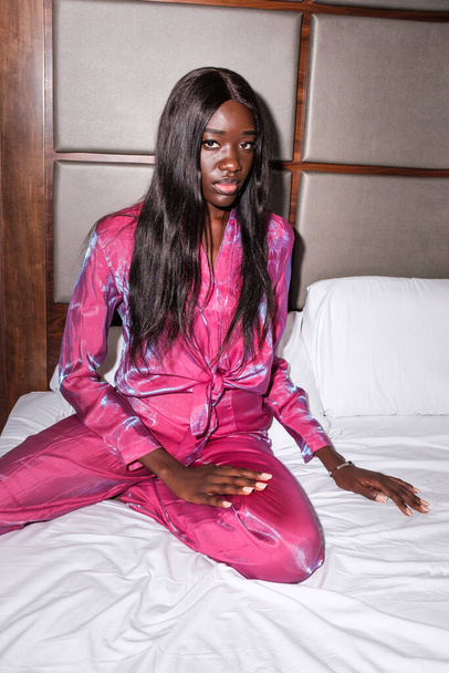 Portrét mladé černošky sedící na hotelovém lůžku. Světlo. Dívá se na kameru a má na sobě okouzlující růžový oblek.. - Fotografie, Obrázek