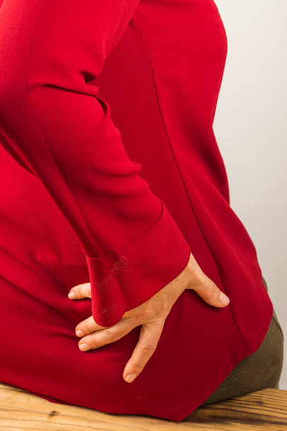 Vertical view of a woman's hand, with a red shirt, holding her waist due to muscle pain, leaning on a table - Photo, Image