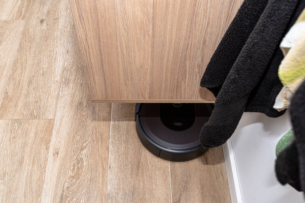 A modern robotic vacuum cleaner cleans the ceramic tiles in the bathroom next to the wall mounted cabinet, an autonomous cleaning robot. - Photo, Image