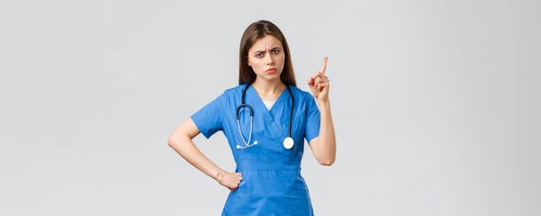 医療従事者、医療、 covid-19と予防接種の概念。青い頭皮で若い女性看護師や医者を流れる,眉をひそめる,患者の悪い行動を叱るとして指を振る,灰色の背景 - 写真・画像