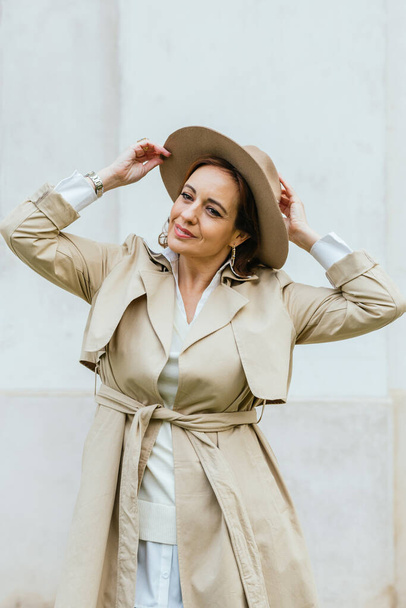 Vrouw in stijlvolle trench jas en hoed op zoek naar camera terwijl poseren buiten op straat. - Foto, afbeelding
