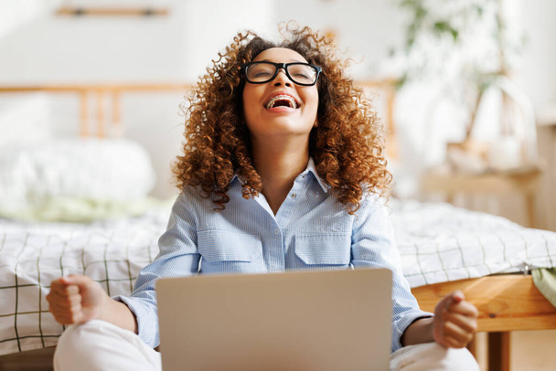 Radosna, szczęśliwa afrykańska nastolatka siedząca w domu w łóżku i patrząca na ekran laptopa z podnieconym wyrazem twarzy, otrzymująca e-mail z dobrymi wiadomościami lub świętująca przyjęcie na uczelnię marzeń. - Zdjęcie, obraz
