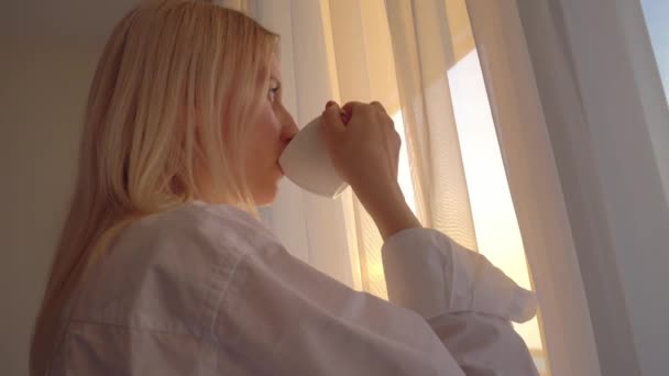 blondi valkoisessa paidassa näyttää ulos ikkunasta, juo kuumaa teetä tai kahvia, - Materiaali, video