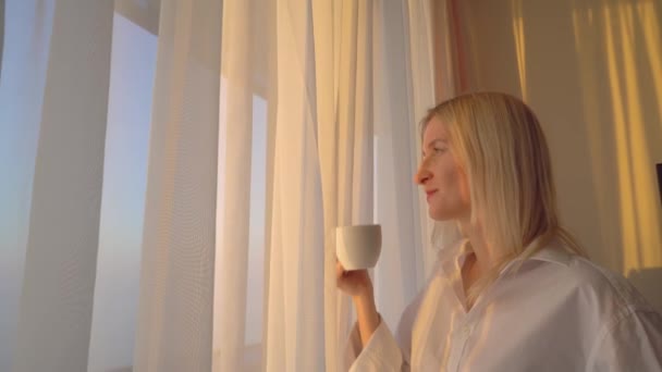 blondynka w białej koszuli wygląda przez okno, pije gorący napój herbaty lub kawy, - Materiał filmowy, wideo