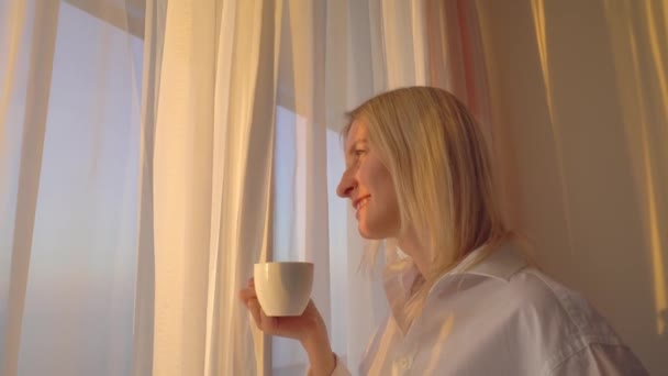 Eine nachdenkliche junge Blondine mit einer Tasse Kaffee oder Tee schaut aus dem Fenster, - Filmmaterial, Video