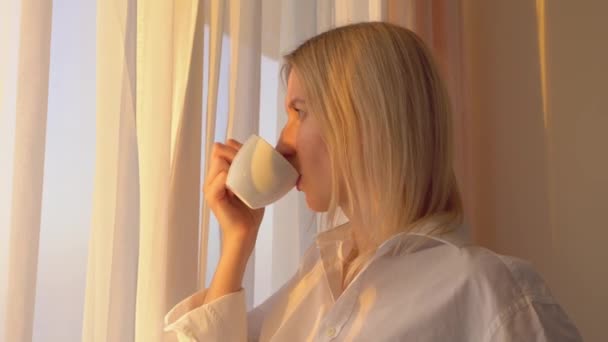 rubia con una camisa blanca mira por la ventana, bebe una bebida caliente de té o café, - Imágenes, Vídeo