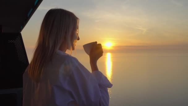 een vrouw bewondert de zonsondergang boven de zee, en drinkt koffie op een hoge verdieping. - Video