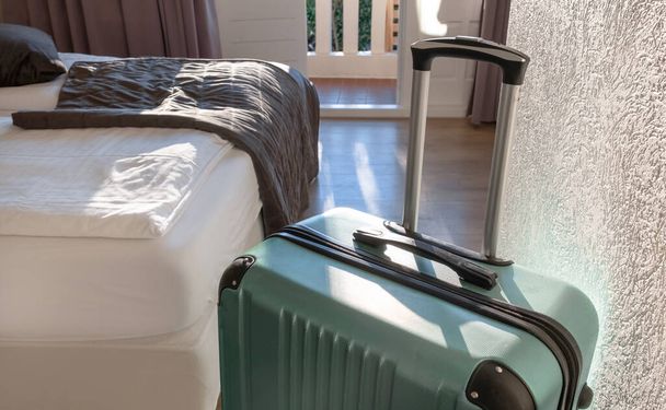 Gran maleta verde se queda en la habitación en el hotel cerca de la ventana o balcón con luz solar. - Foto, Imagen