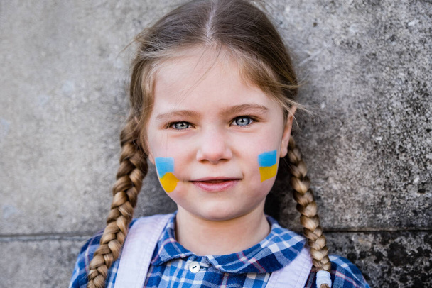 ウクライナの国旗の黄色-青の色で頬に描かれた旗を持つおびえた子供の少女の肖像画。平和と保護の子供の概念 - 写真・画像