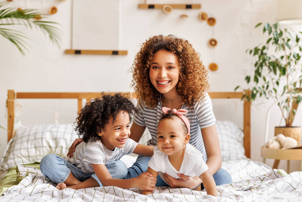 happy ethnic curly mom playing with laughing little son and daughter in bed, playful little children merrily looking at mother - Foto, Imagen