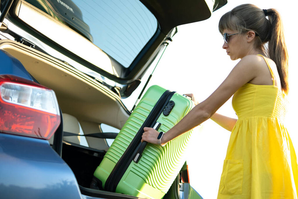 Nuori naiskuljettaja ottaa vihreän matkalaukun auton takakontista. Matkailu ja lomat käsite - Valokuva, kuva
