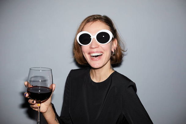 Retrato de una joven despreocupada festejando y sosteniendo una copa de vino mientras usa gafas de sol, disparada con flash - Foto, Imagen