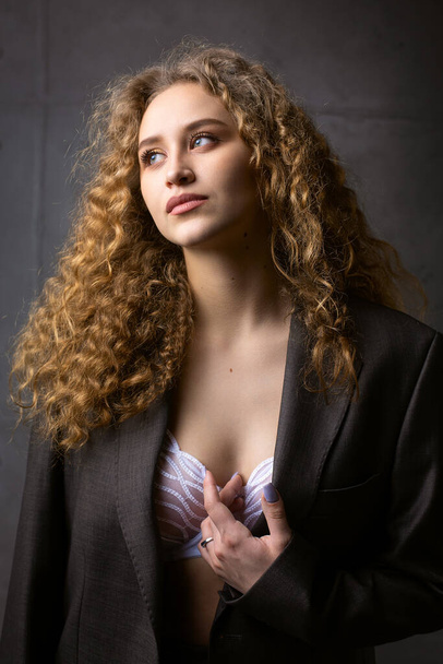 Πορτρέτο ενός νεαρού κοκκινομάλλη σγουρομάλλη σέξι κορίτσι σε ένα σακάκι με μακριά μαλλιά στο στούντιο. Δραματική φωτογραφία σε σκούρα χρώματα. - Φωτογραφία, εικόνα