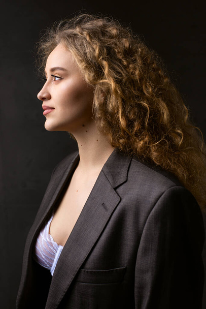 Portret van een jong roodharig gekruld sexy meisje in een jasje met lang haar in de studio. Dramatische foto in donkere kleuren. - Foto, afbeelding