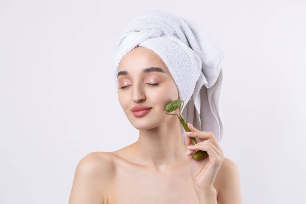 Beautiful girl with thick eyebrows and perfect skin at white background, towel on head, beauty photo. Holds a cosmetic roller. - Foto, Bild