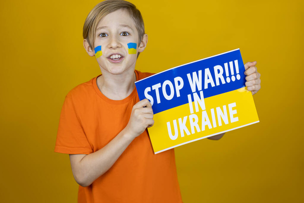 ένα αγόρι με μια ζωγραφισμένη σημαία της Ουκρανίας στα μάγουλά του κρατά μια αφίσα στο πλάι με την επιγραφή σταματήσει τον πόλεμο στην Ουκρανία και φώναξε - Φωτογραφία, εικόνα