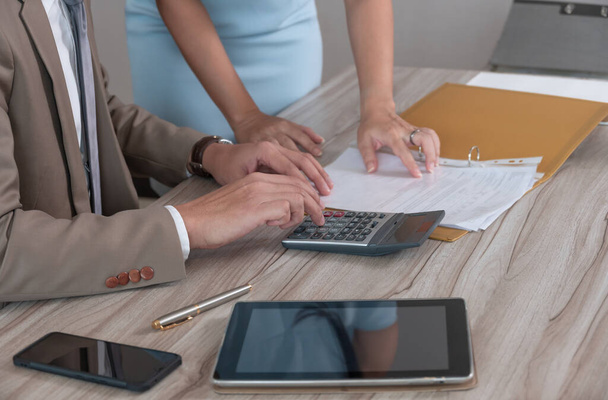 Businessman manager in brown suit using calculator while sitting at office wooden desk and discussing on business document with female colleague in blue dress. Business analyst, investment, accounting, finance and  teamwork concept. - Photo, Image
