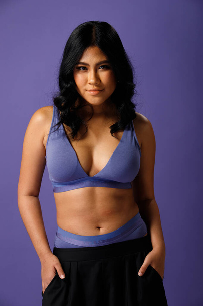 Asian 20s Woman chubby wear sport bra in purple color or Very Peri world trend over purple background. Half body Female black hair express feeling strong smile on face eyes in studio lighting - Photo, Image