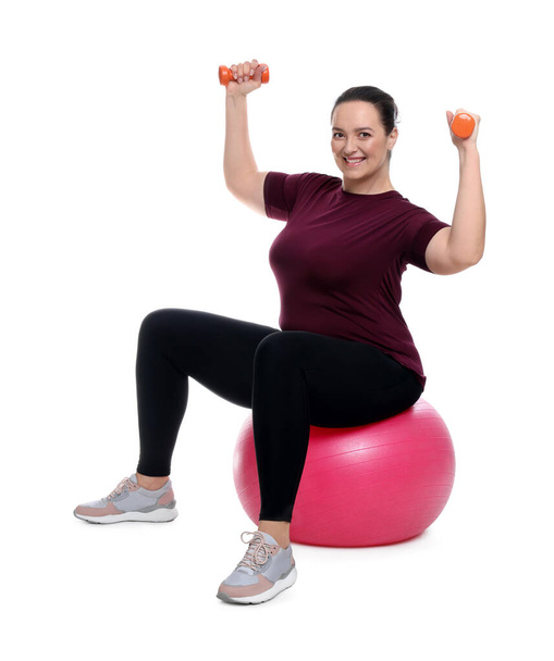 Happy overweight woman with dumbbells sitting on fitness ball against white background - Foto, immagini