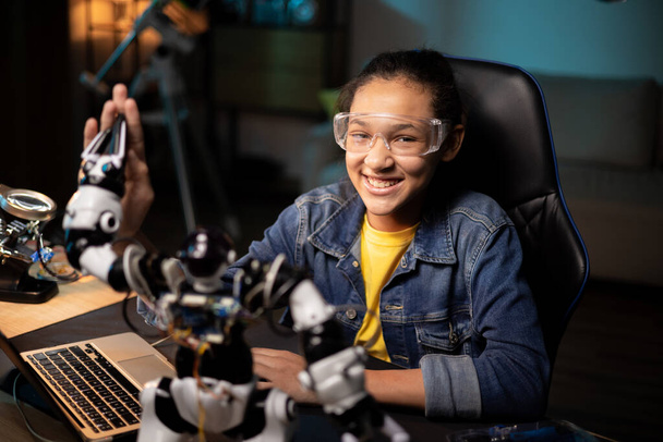 Портрет улыбающейся девушки, проводящей вечер за столом, проверяющей инструкции по игрушкам на ноутбуке, просматривающей веб-страницы, вытягивающей электронные кабели от робота, фиксирующей - Фото, изображение