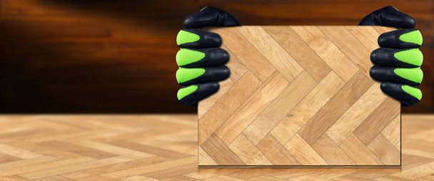 Dvě ruce s ochrannými pracovními rukavicemi, které drží vzorek parkety z herringového dřeva na podobné podlaze, s kopírovacím prostorem. - Fotografie, Obrázek