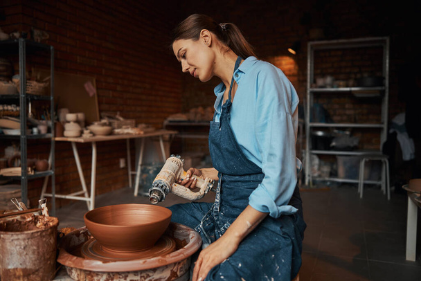 Brunette female artisan modeling ceramic tableware on special equipment in pottery workshop - Photo, Image