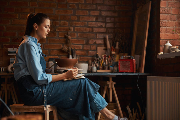 Happy positive thoughtful Caucasian craft woman writing something in pottery workshop - 写真・画像