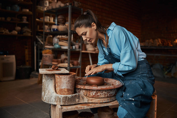 Beautiful elegant craft woman working with fresh wet clay on special equipment in pottery workshop - Фото, зображення