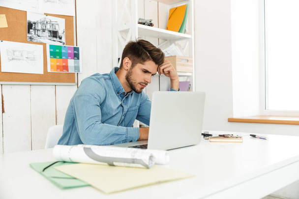Young white man working with laptop while sitting at desk in office indoors - Foto, Imagen