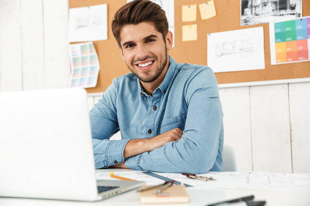 Young white man smiling while working with laptop and drawings in office indoors - Foto, Imagem