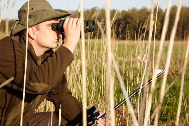 Después de una inspección más cercana. Un hombre en la vida salvaje mirando a través de sus prismáticos sosteniendo su rifle. - Foto, imagen