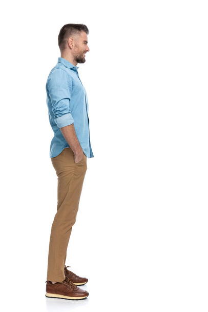 side view of happy bearded guy with hands in pockets standing in line and smiling while waiting in a queue on white background in studio - Foto, Bild