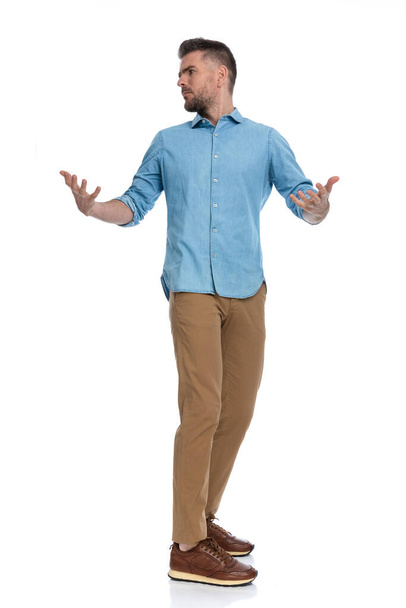 hombre casual en camisa de mezclilla mirando detrás del hombro y abriendo los brazos como un gesto nervioso mientras está de pie en línea sobre fondo blanco - Foto, Imagen
