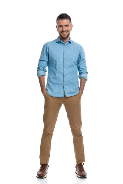 homem casual feliz com as mãos nos bolsos vestindo camisa jeans azul, calças de chino e tênis e sorrindo no fundo branco, corpo inteiro - Foto, Imagem