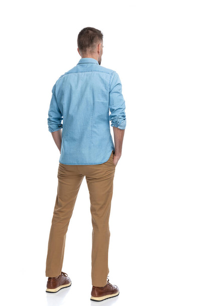 behind view of casual young guy wearing denim shirt, chino pants and sneakers holding hands in pockets and looking away on white background - Photo, Image