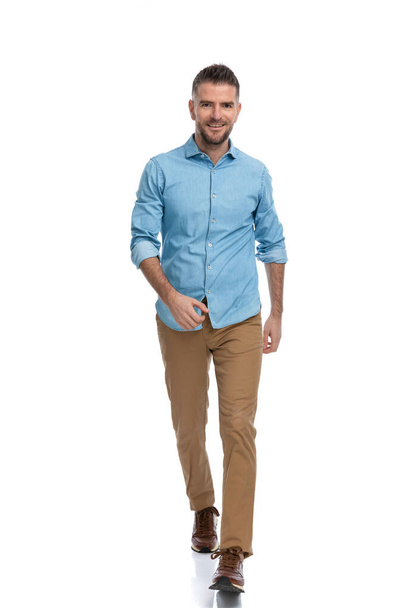 full body picture of confident unshaved man in casual outfit on white background walking and smiling in studio - Photo, Image